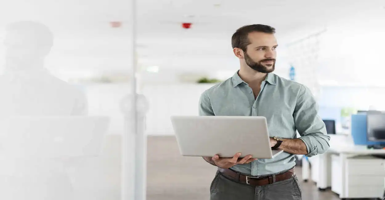 Homem em pé com um notebook na mão dentro de uma empresa